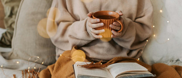  A person on a couch covered by a blanket, holding a mug of tea and reading a bo